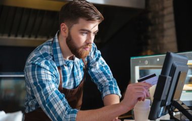 Handsomeconcentratedbeardedwaiterswipingcreditcardthroughthecomputerterminalincafe