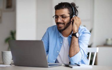 Smiling Young Indian Guy Watching Webinar On Laptop, Wearing Headset And Eyeglasses, Enjoying Online Education And Distance Learning, Sitting At Desk At Home, Looking At Computer Screen, Free Space