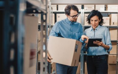 Female Inventory Manager Shows Digital Tablet Information to a Worker Holding Cardboard Box, They Talk and Do Work. In the Background Stock of Parcels with Products Ready for Shipment.