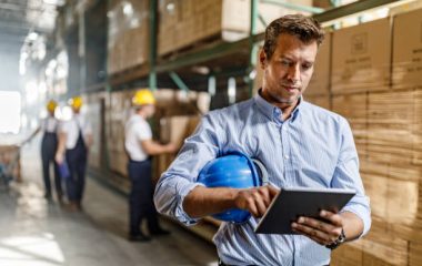 Mid adult foreman surfing the Internet on touchpad in a distribution warehouse. There are people in the background.
